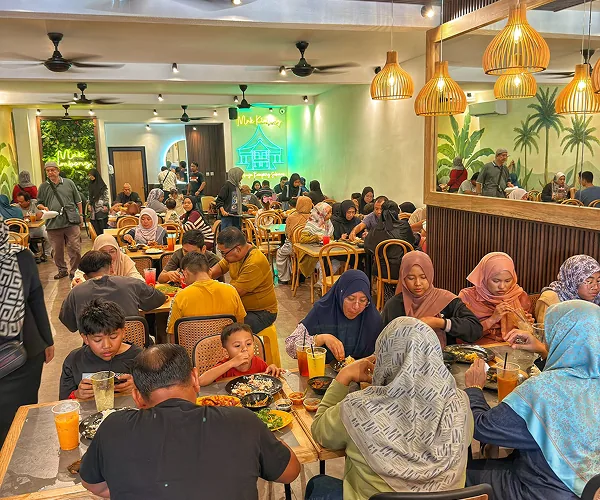 Ruang makan Restoran Mak Kimbong yang penuh dengan keluarga makan bersama, di bawah lampu rotan dan mural pokok kelapa