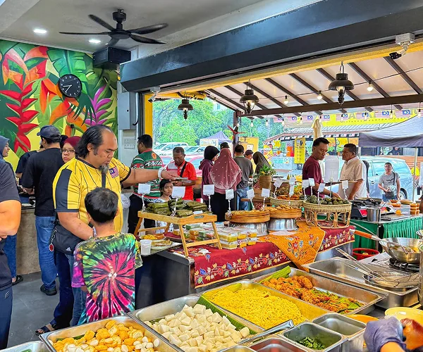 Kaunter depan Restoran Mak Kimbong: nasi impit, mi dan kuih di atas meja berlapik batik, dengan pelanggan di sekelilingnya