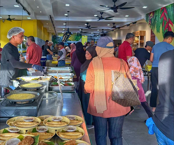 Tukang masak berpenutup rambut di dapur kaunter Restoran Mak Kimbong, pelanggan beratur, dan pinggan hidangan berlipat dengan mangkuk kecil kuah di hadapan